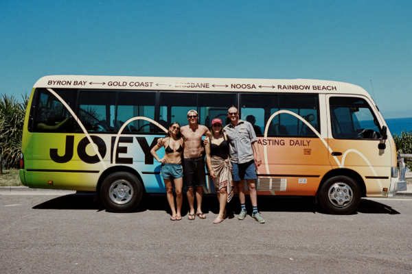 A group of friends standing in front of the multicoloured JOEY bus with the ocean in the background