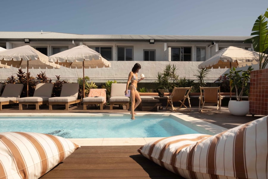 A girl with a coconut dipping her toes into the water of a rooftop swimming pool surronded by sun loungers