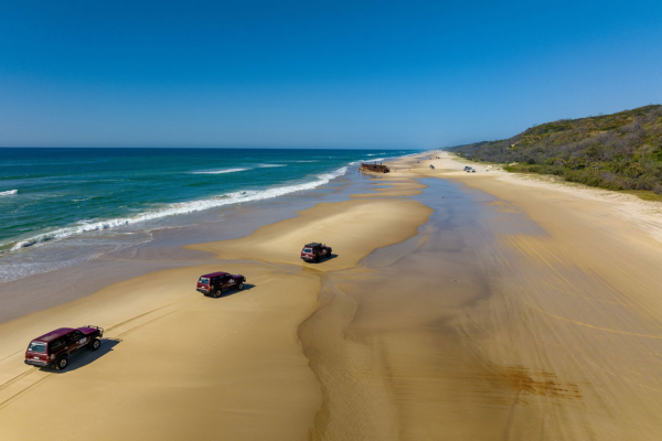 Three vehicles driving in a convoy along 75 mile beach with a shipwreck on the beach and ocean on one side and forest on the other side
